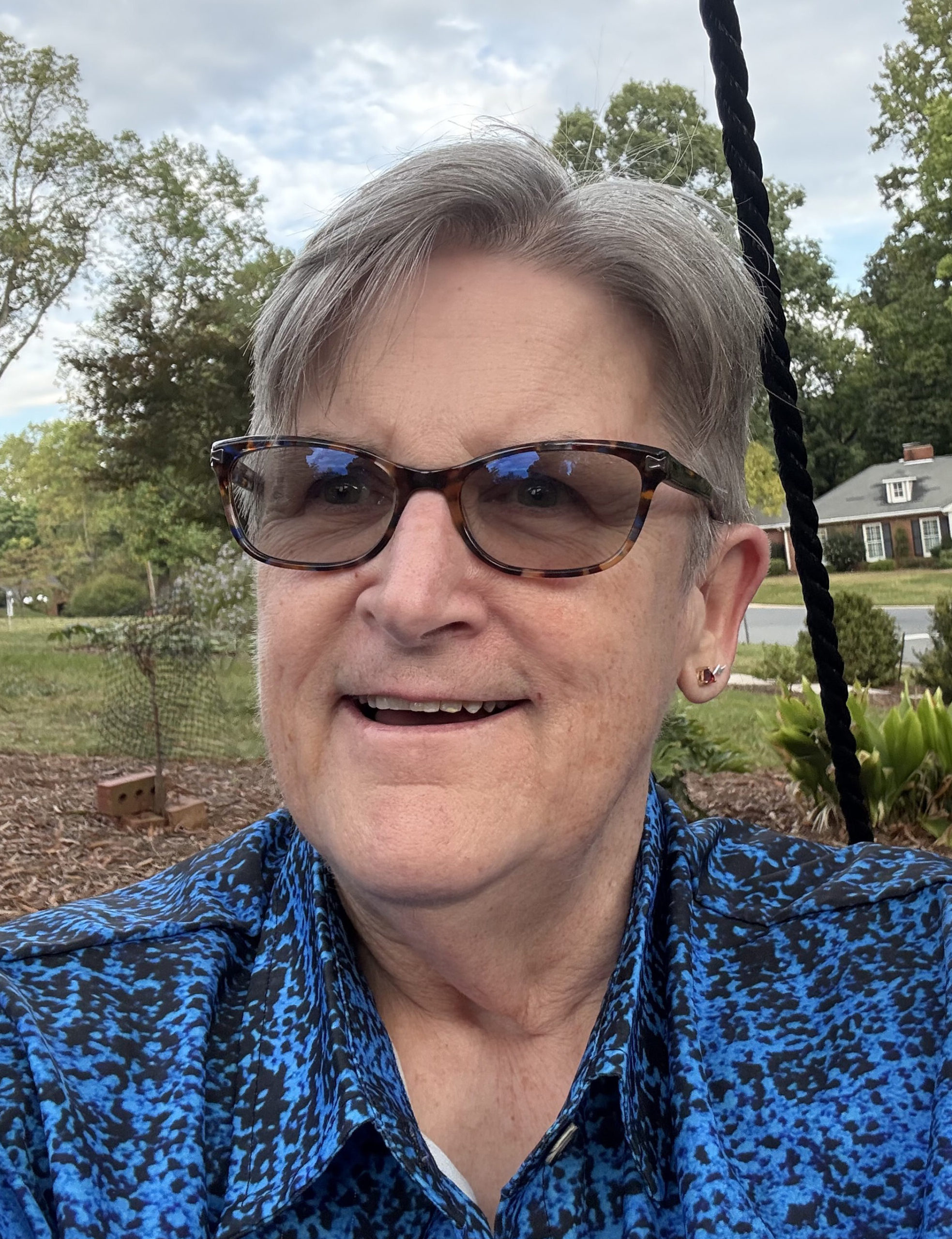 An older woman wearing sunglasses and a blue shirt smiles at the camera, exuding a relaxed and cheerful demeanor.