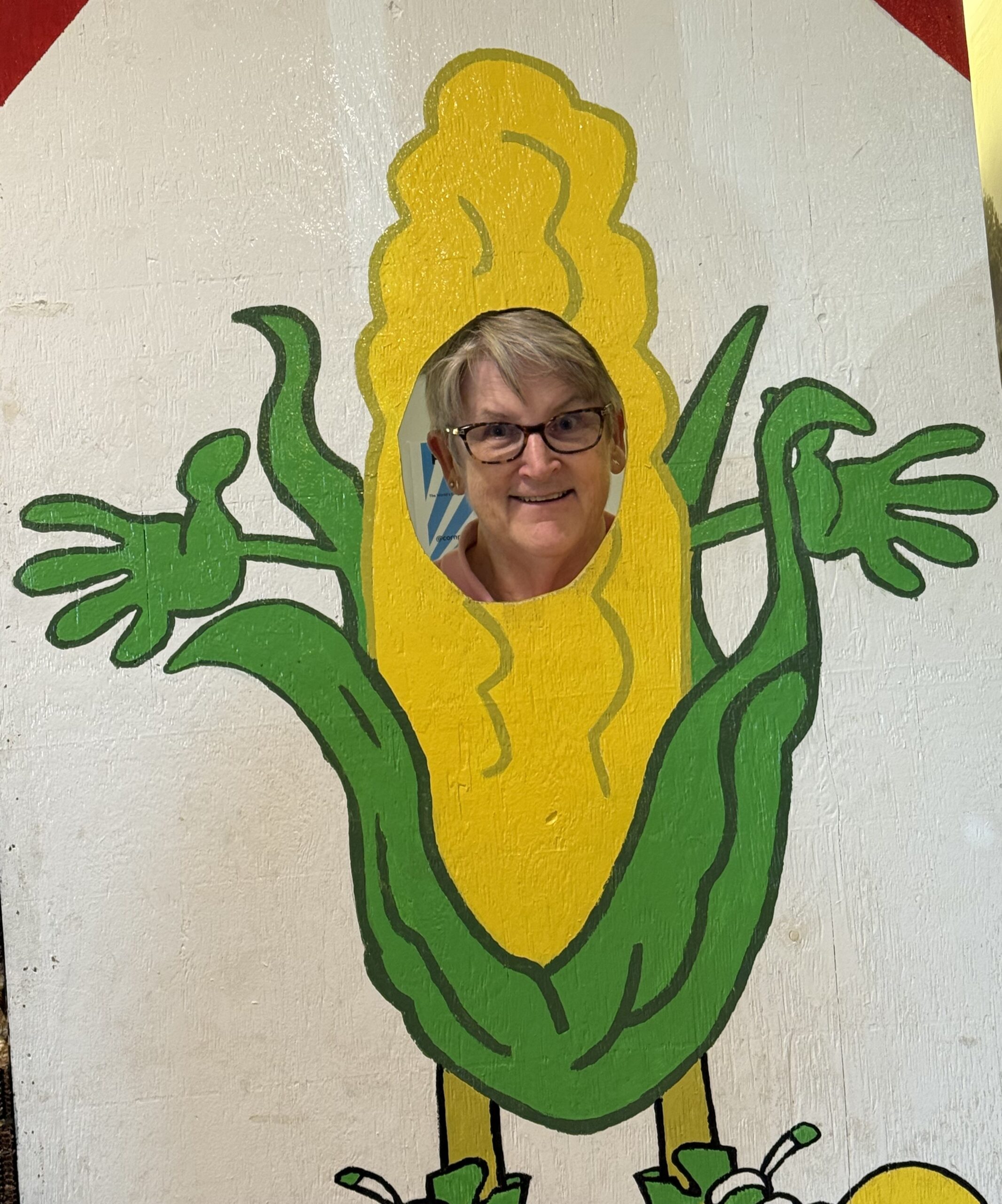 A woman dressed in a corn costume stands in front of a colorful sign, smiling and posing for the camera.