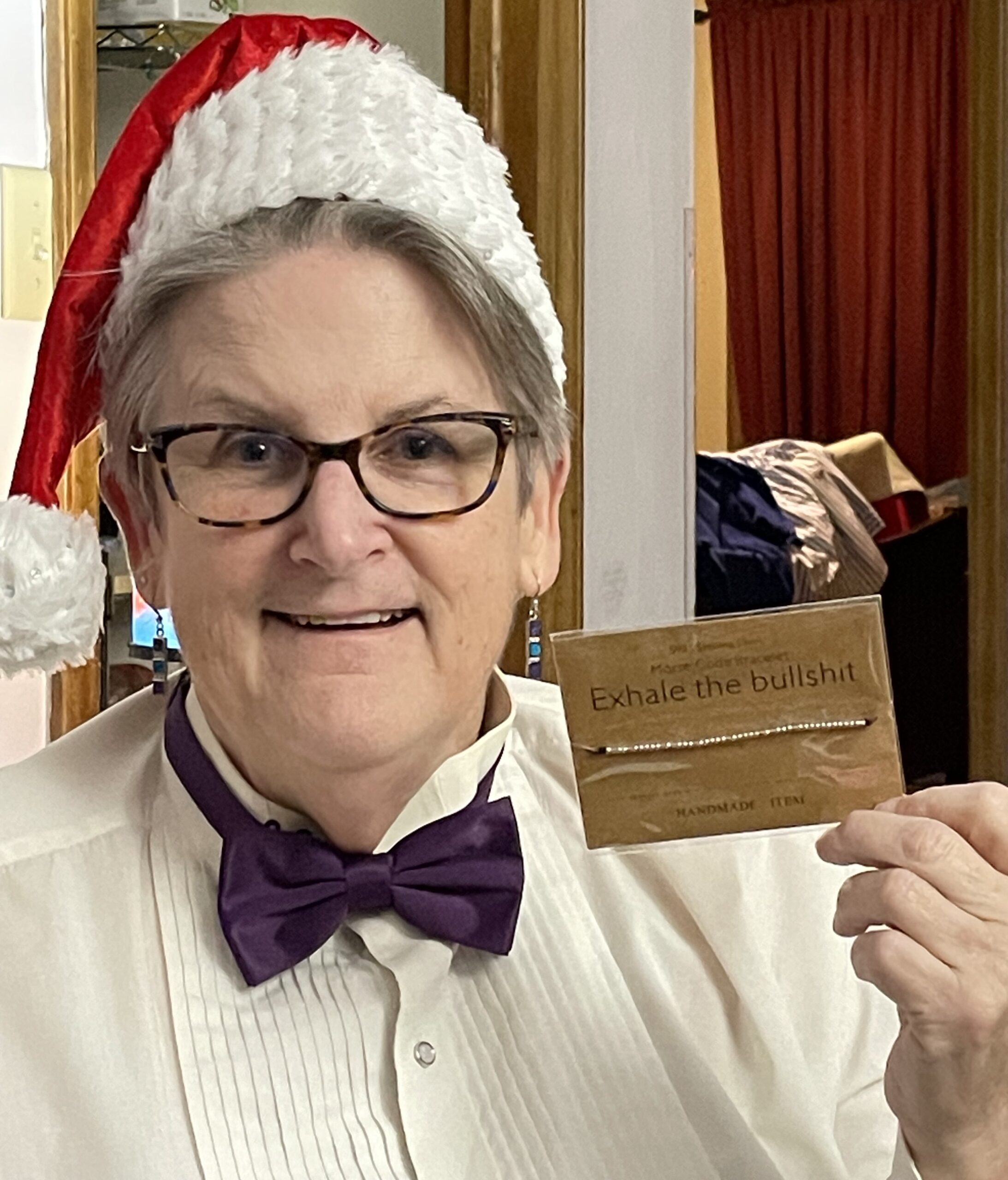 A woman in a Santa hat and glasses holds a wooden card, smiling warmly at the camera.
