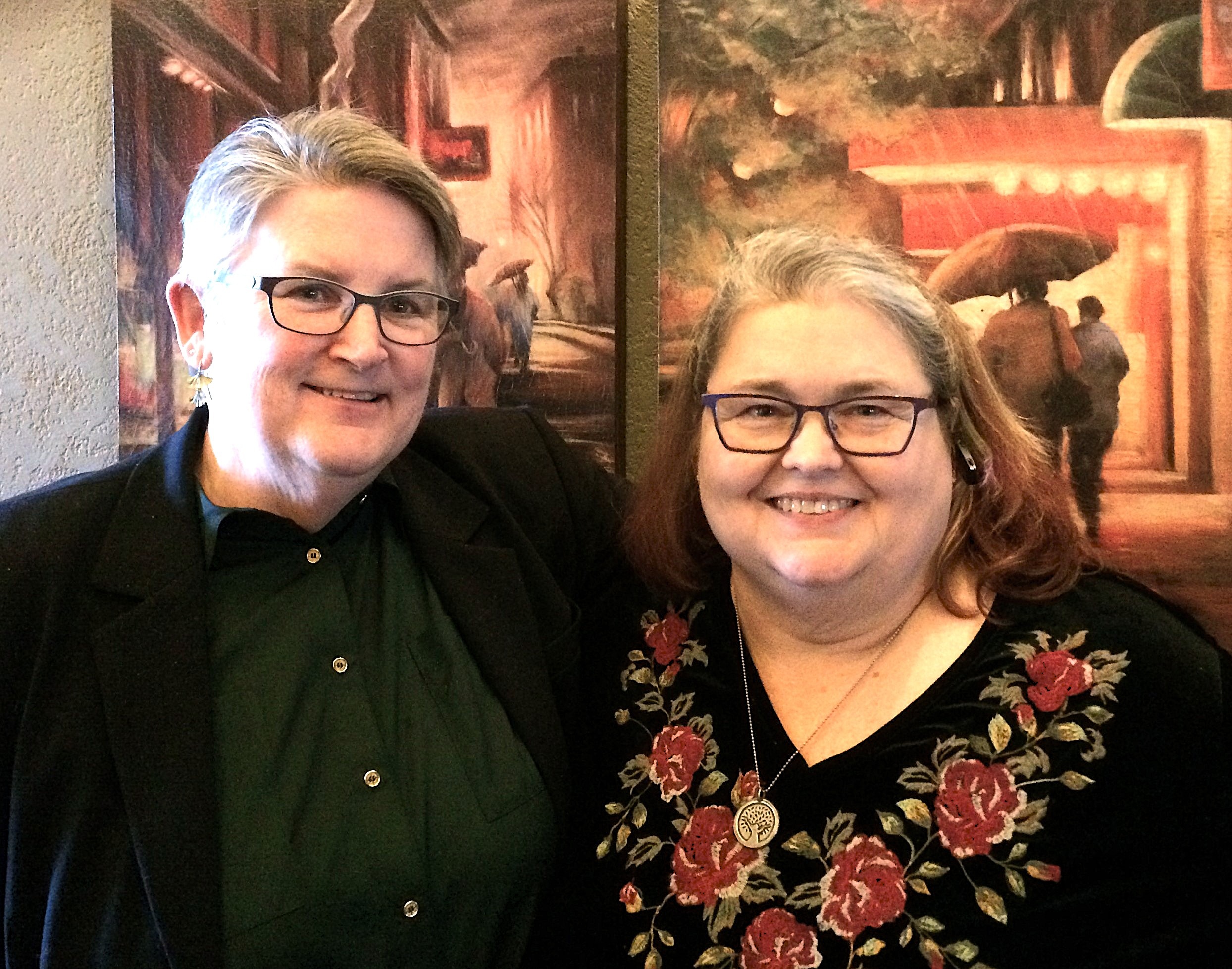 Two women stand side by side, smiling in front of colorful paintings in an art gallery.