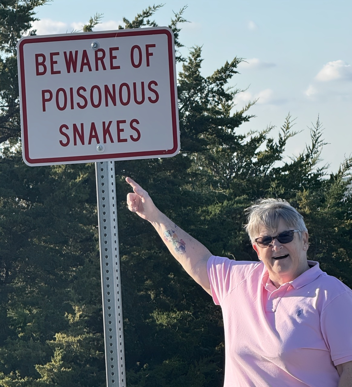 A woman points at a warning sign that reads "Beware of Poisonous Snakes" in a natural outdoor setting.